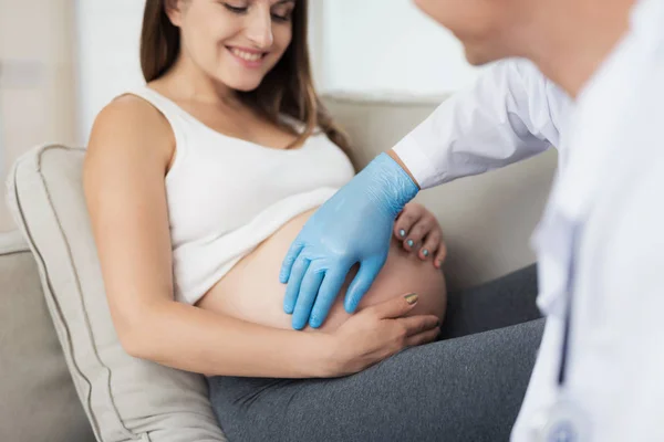 A pregnant woman lies on a light sofa at home. A doctor came to her, he probes her belly — Stock Photo, Image