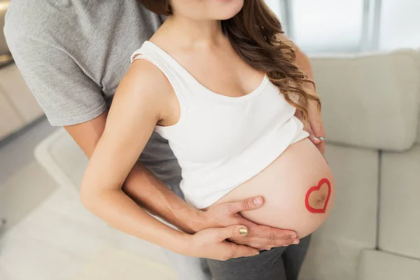 Nahaufnahme. Eine schwangere Frau posiert mit ihrem Mann. Er umarmt sie von hinten. Sie hat ein Herz auf dem Bauch — Stockfoto