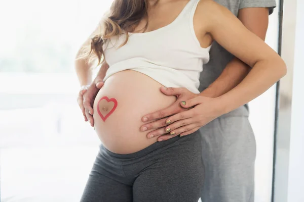 Nahaufnahme. Eine schwangere Frau posiert mit ihrem Mann. Er umarmt sie von hinten. Sie hat ein Herz auf dem Bauch — Stockfoto