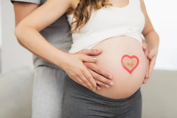 Fecha. Uma mulher grávida posando com o marido. Ele abraça-a por trás. Ela tem um coração na barriga. — Fotografia de Stock