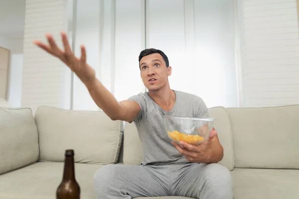 Mężczyzna w szarym domu ubrania siedzi na kanapie i odpoczynku. On entuzjastycznie ogląda telewizję i pije piwo — Zdjęcie stockowe