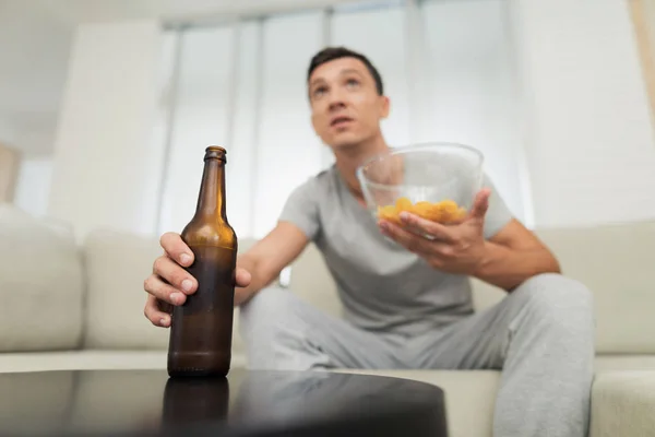 Un hombre con ropa de casa gris está sentado en el sofá y descansando. Ve con entusiasmo la televisión y bebe cerveza. —  Fotos de Stock