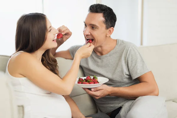 Una mujer embarazada yace en un sofá ligero. Un hombre está sentado a su lado. Comen fresas y dulces. —  Fotos de Stock