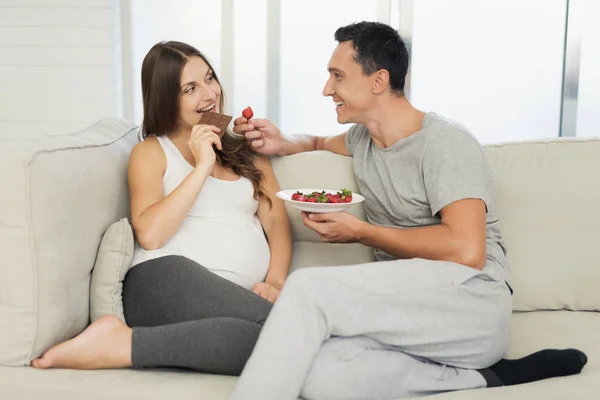 Беременная женщина лежит на светлом диване. Рядом с ней сидит мужчина. Они едят клубнику и сладости. — стоковое фото
