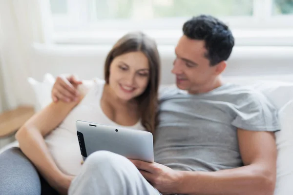 Una donna incinta e un uomo sono sdraiati sul letto. Stanno guardando qualcosa sul loro tablet — Foto Stock