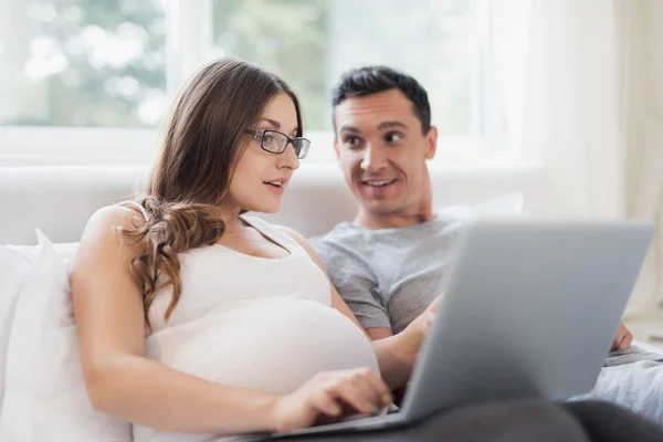 Eine schwangere Frau und ein Mann liegen auf dem Bett. eine Frau hält einen Laptop auf ihrem Schoß, ein Mann hält ein Smartphone. — Stockfoto