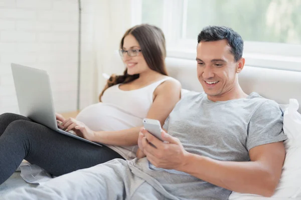 A pregnant woman and a man are lying on the bed. A woman is holding a laptop on her lap, a man is holding a smartphone. — Stock Photo, Image