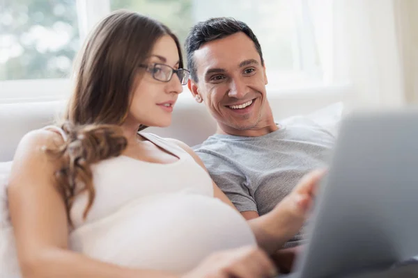 Eine schwangere Frau und ein Mann liegen auf dem Bett. eine Frau hält einen Laptop auf ihrem Schoß, ein Mann hält ein Smartphone. — Stockfoto