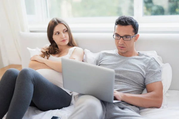 Eine schwangere Frau und ein Mann liegen auf dem Bett. ein Mann hält einen Laptop auf seinem Schoß und arbeitet — Stockfoto