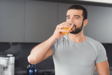 Sportif bir adam mutfakta duruyor ve portakal suyu içiyor. Onun yanında bir yığın sebze ve meyve üzerinde.
