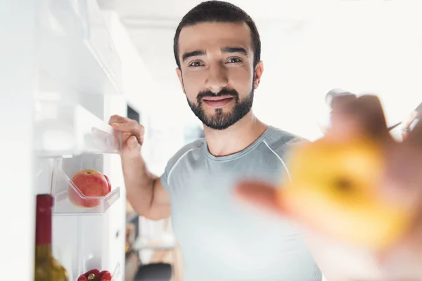Un deportista se para en la cocina y toma verduras frescas del refrigerador . —  Fotos de Stock