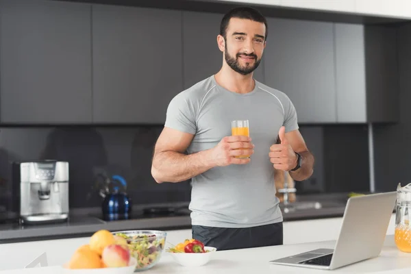 Um homem esportivo está posando na cozinha. Ele está segurando um copo de suco e mostrando seu polegar para cima . — Fotografia de Stock