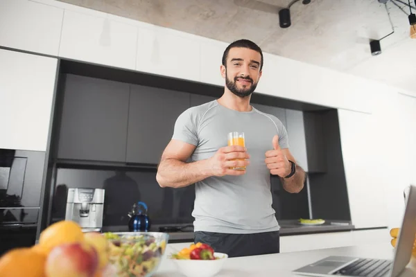 Um homem esportivo está posando na cozinha. Ele está segurando um copo de suco e mostrando seu polegar para cima . — Fotografia de Stock