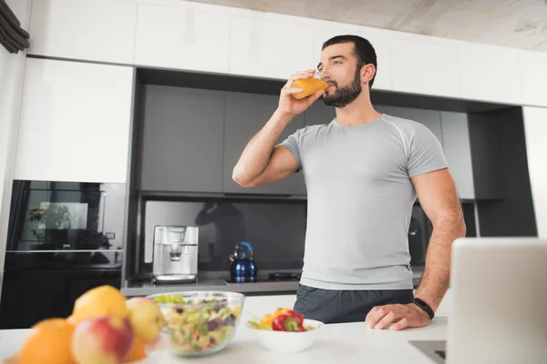 Un deportista se para en la cocina y bebe jugo de naranja. Junto a él en una pila de verduras y frutas . —  Fotos de Stock