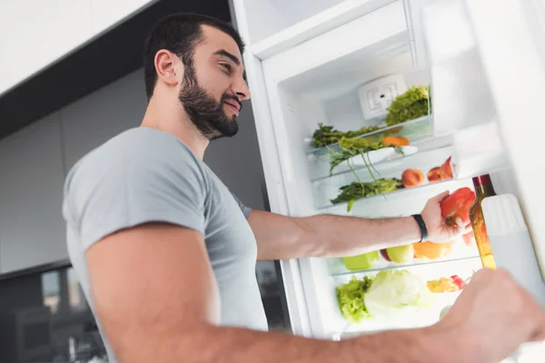 Un deportista se para en la cocina y toma verduras frescas del refrigerador . — Foto de Stock