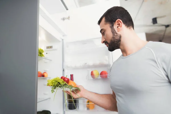 Um homem desportivo fica na cozinha e tira legumes frescos do frigorífico . — Fotografia de Stock
