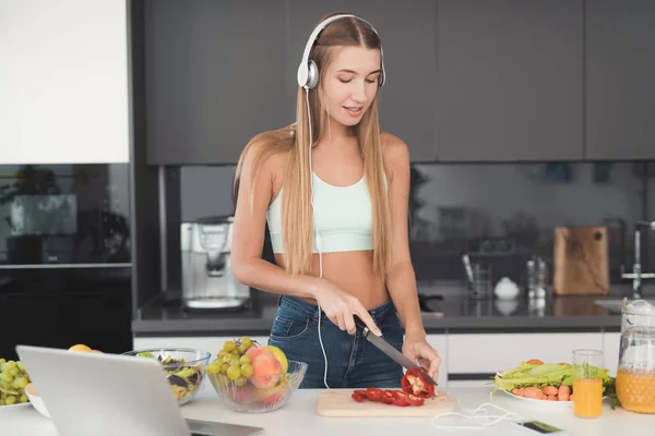 Молодая спортсменка стоит на кухне и готовит салат. У неё на голове белые наушники. . — стоковое фото