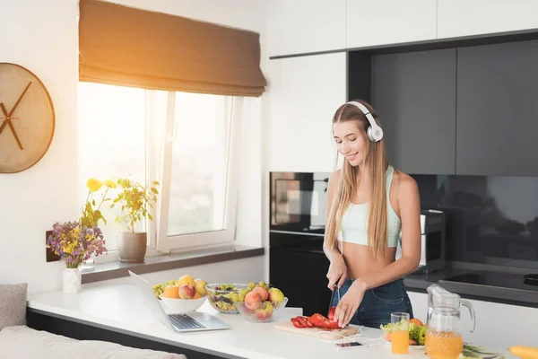 Chica atlética joven está de pie en la cocina y está preparando una ensalada. Tiene auriculares blancos en la cabeza. . —  Fotos de Stock