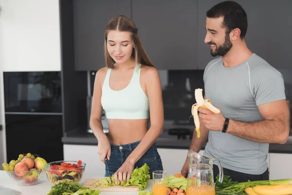 Een paar bereidt een salade. Een meisje is de groenten snijden voor een salade. Mens is permanent volgende en eten van een banaan. — Stockfoto