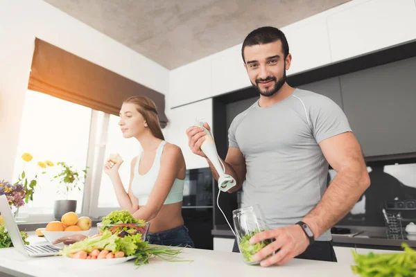 Jonge sport paar in de ochtend in de keuken bereidt. Een man maakt een groene groente cocktail. — Stockfoto