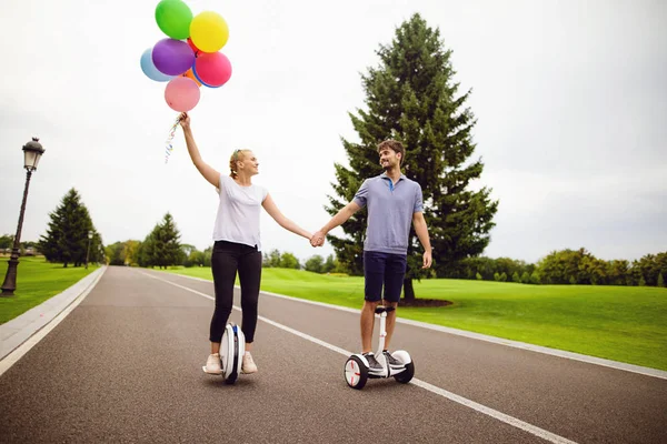 La pareja está montando un giroboard y un monóculo en el parque. Están contentos. Mujer sosteniendo globos en las manos — Foto de Stock