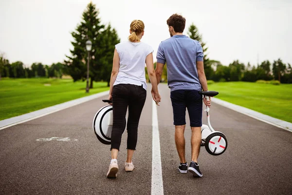 Um homem e uma mulher caminham pelo caminho do parque. Eles dão as mãos e carregam o seu monocluto e giroscópio em suas mãos — Fotografia de Stock