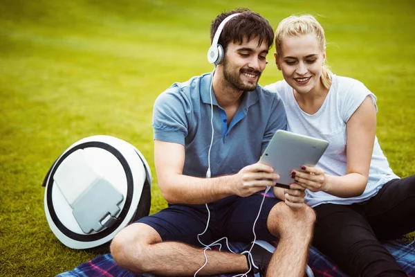 A couple is resting in the park after a walk. Nearby is a monoclean. They are holding a tablet. — Stock Photo, Image