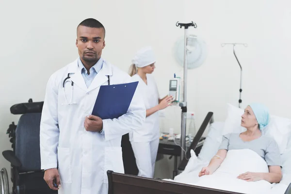 Läkare poserar mot bakgrund av patienten. Han innehar en papper-tablett i händerna. — Stockfoto