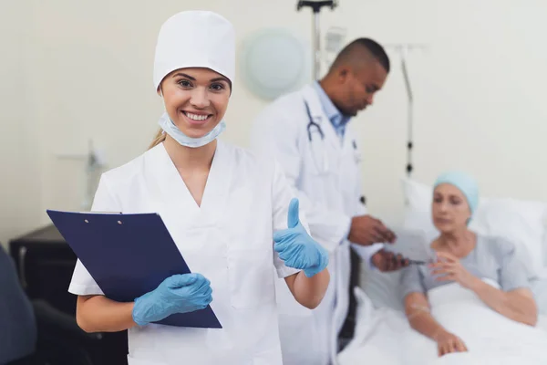 En sjuksköterska poserar mot patientens bakgrund. Hon håller en papper-tablett i händerna. — Stockfoto