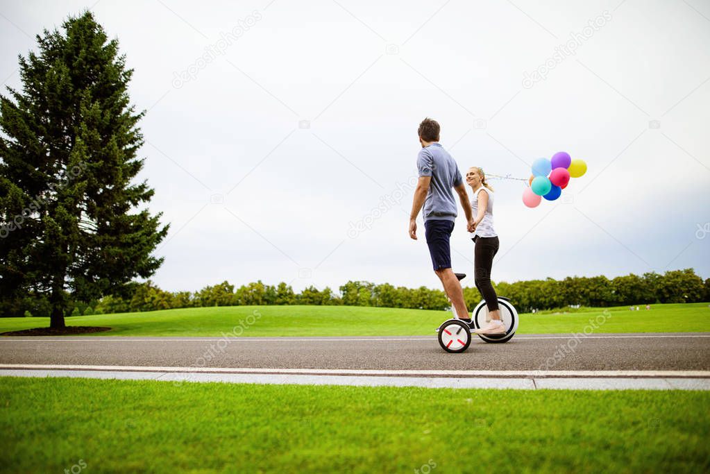 The couple is riding a gyroboard and a monocle in the park. They are happyy. A woman holds balloons in her hands