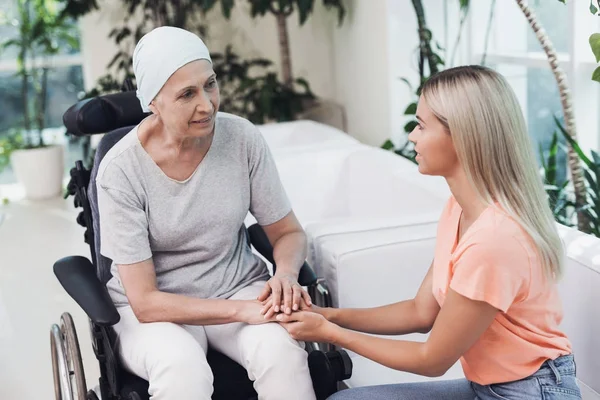 Una mujer con cáncer está sentada en una silla de ruedas. Junto a ella se sienta su hija en un sofá blanco . —  Fotos de Stock