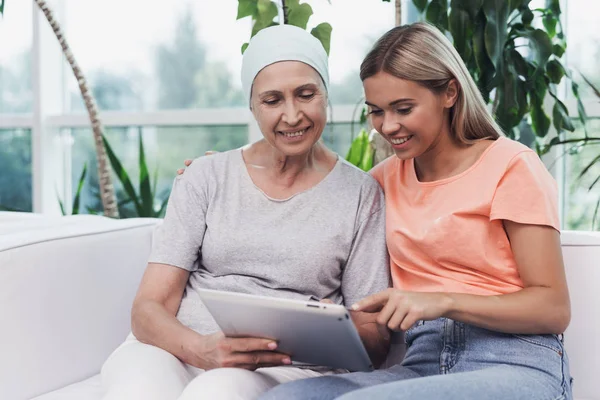 Una mujer con cáncer está sentada junto a su hija en un sofá blanco. Están mirando algo en una tableta gris . —  Fotos de Stock