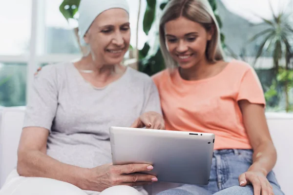 Una mujer con cáncer está sentada junto a su hija en un sofá blanco. Están mirando algo en una tableta gris . — Foto de Stock