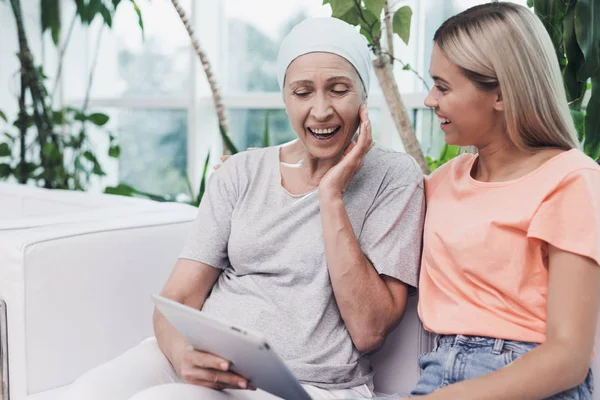 Una mujer con cáncer está sentada junto a su hija en un sofá blanco. Están mirando algo en una tableta gris . —  Fotos de Stock