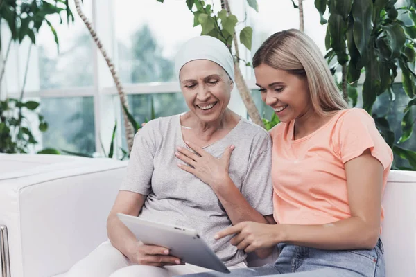 Una mujer con cáncer está sentada junto a su hija en un sofá blanco. Están mirando algo en una tableta gris . —  Fotos de Stock