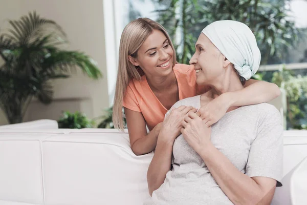 Жінка з раком сидить на білому дивані поруч зі своєю дочкою. Дівчина обіймає жінку — стокове фото