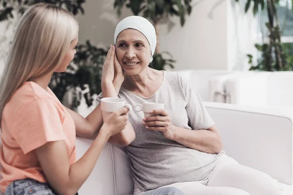 Una mujer con cáncer está sentada en un sofá blanco junto a su hija. Se sientan con tazas de té en sus manos . —  Fotos de Stock