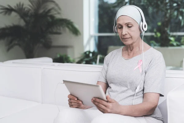 Una mujer con cáncer está sentada en un sofá blanco en una clínica moderna. Tiene una cinta rosa en la camisa. . —  Fotos de Stock
