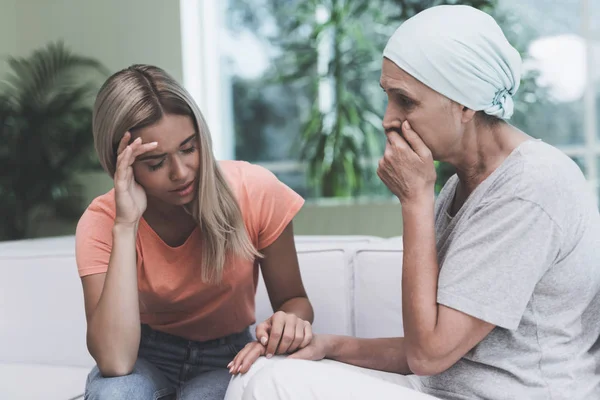 A woman with cancer is sitting on a white sofa in a modern clinic. Next to her sits her daughter.