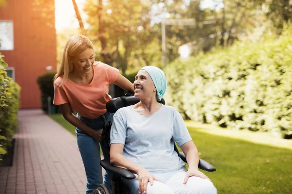 Una mujer con cáncer está sentada en una silla de ruedas. Ella camina por la calle con su hija y se están divirtiendo . —  Fotos de Stock