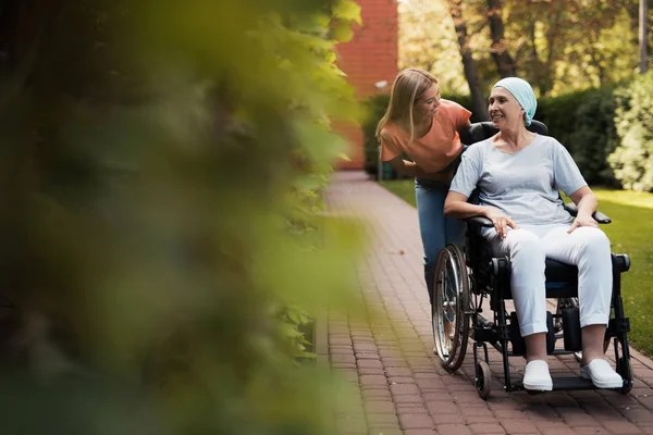 Uma mulher com cancro está sentada numa cadeira de rodas. Ela caminha na rua com sua filha e eles estão se divertindo . — Fotografia de Stock