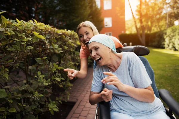 Uma mulher com cancro está sentada numa cadeira de rodas. Ela caminha na rua com sua filha e eles estão se divertindo . — Fotografia de Stock