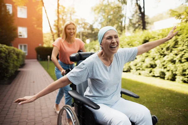 Uma mulher com cancro está sentada numa cadeira de rodas. Ela caminha na rua com sua filha e eles brincam . — Fotografia de Stock