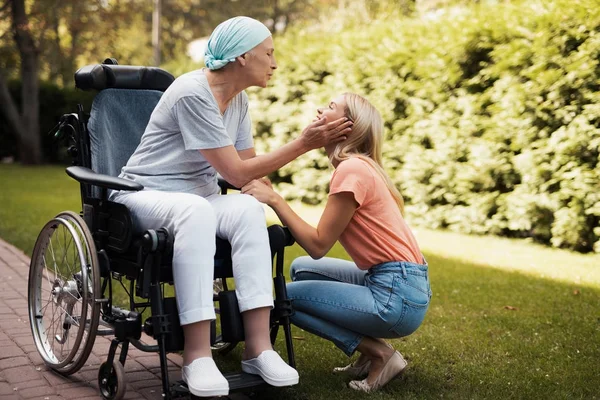 Una mujer con cáncer está sentada en una silla de ruedas. Ella sonríe y sostiene las mejillas de su hija . —  Fotos de Stock