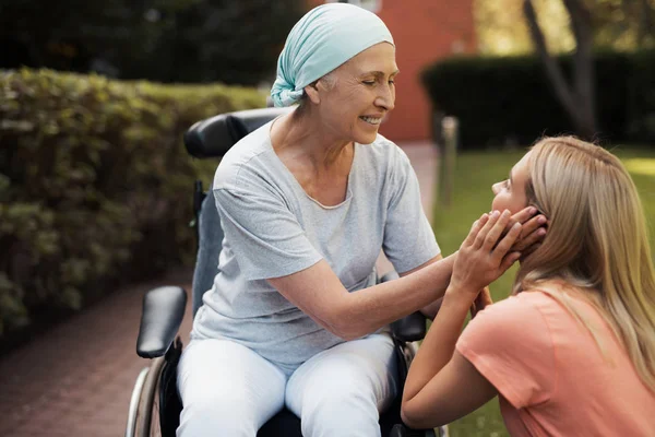 Una mujer con cáncer está sentada en una silla de ruedas. Ella sonríe y sostiene las mejillas de su hija . —  Fotos de Stock