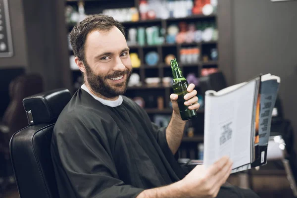 Člověk pije alkohol a čtení Kadeřnické křeslo moderní holičství. — Stock fotografie