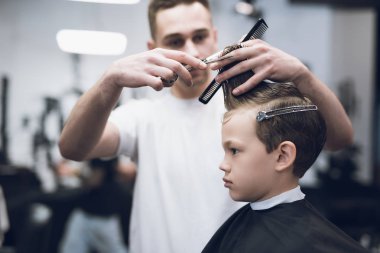 The hairdresser makes a fashionable pretty hairstyle for the boy in a modern barbershop. clipart