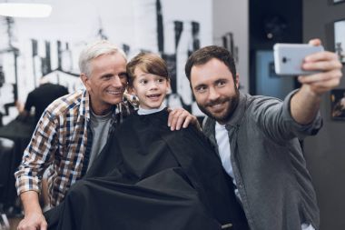 Adult man makes selfie on a smartphone with older men and boy in barbershop. clipart