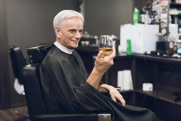 The old man drinks alcohol in the barber\'s chair in barbershop.