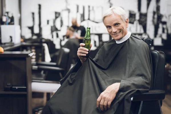 Den gamla mannen dricker alkohol i barberarens stol i barbershop. — Stockfoto